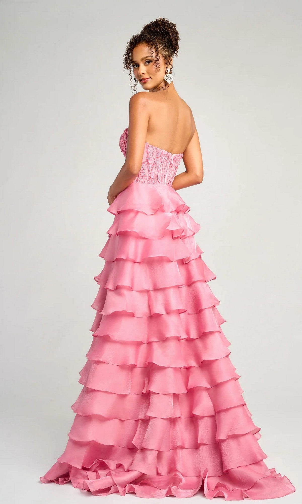 A woman models the Long Formal Dress: Portia and Scarlett PS26048, a strapless, floor-length pink ruffled gown with an embroidered corset bodice, standing against a plain light background and looking over her shoulder.